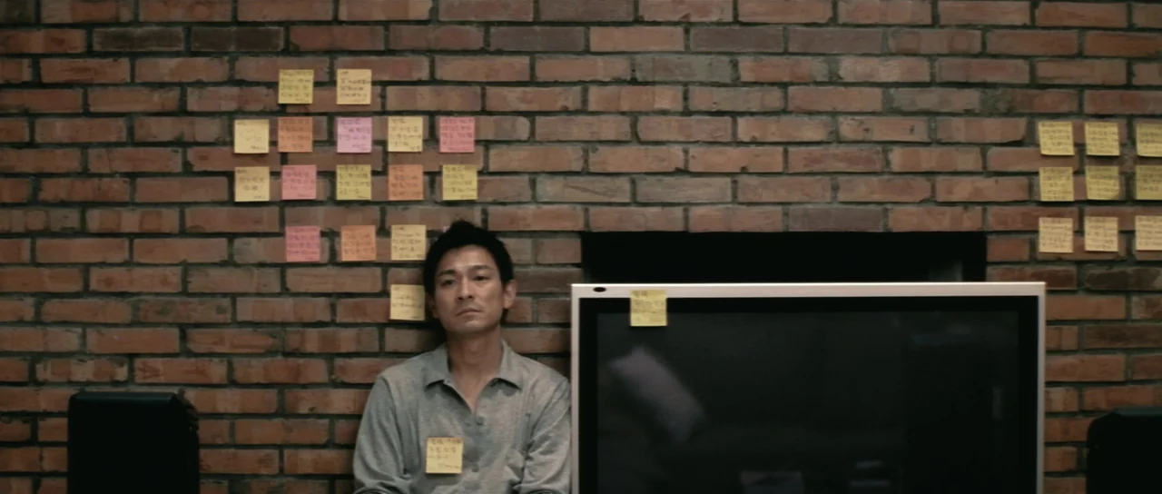 Infernal Affairs (2002) movie still: medium — A man sits against a brown brick wall covered in dozens of small yellow sticky notes. He remains…