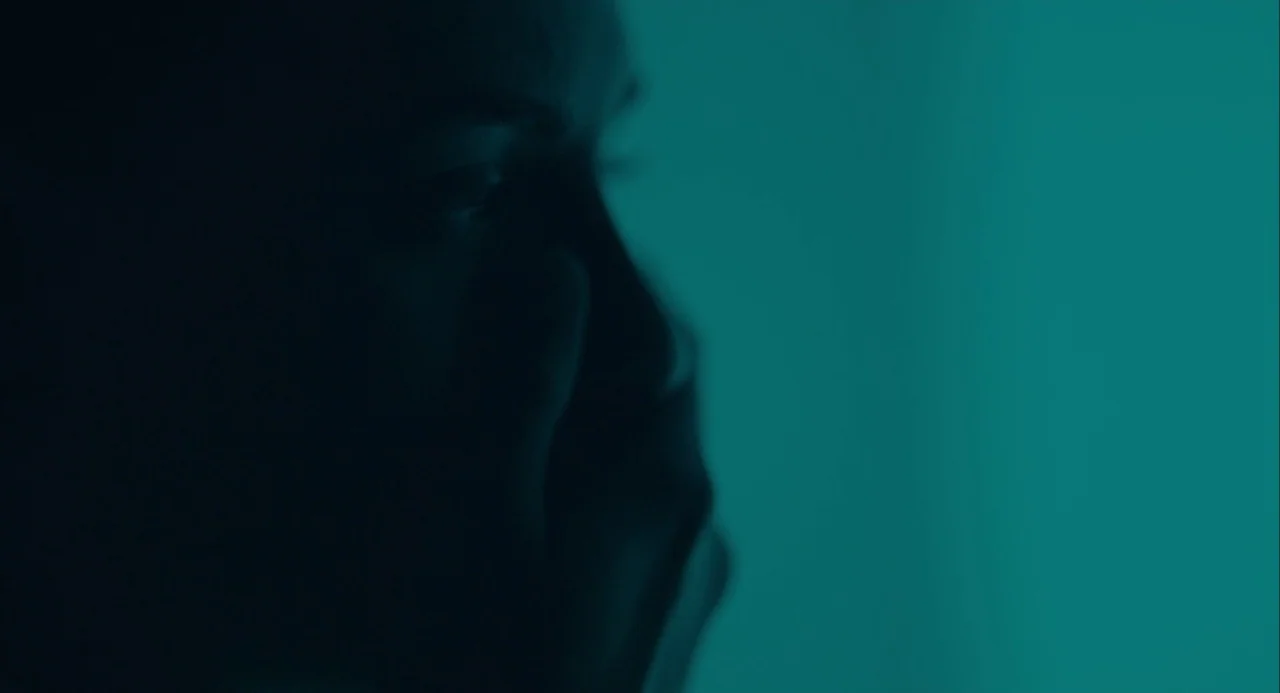Equals (2015) movie still: closeup — A dark space lit by a single cyan light source. A male profile rests his hand on his chin.