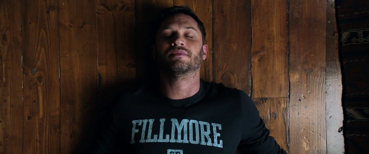 Venom (2018) movie still: medium — A person lies on a polished plank floor made of dark wood. The man is lying flat on his back with his eyes closed.