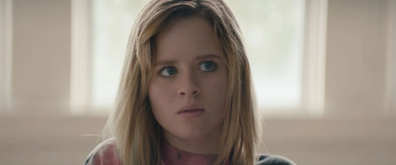 Becky (2020) movie still: closeup — A person is centered in a brightly lit indoor space with a window backdrop. The individual stands still looking…