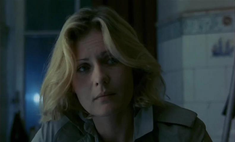 No End (1985) movie still: closeup — A dimly lit room with blue-toned light reflecting off white tiled walls. A woman with blonde hair stares into…