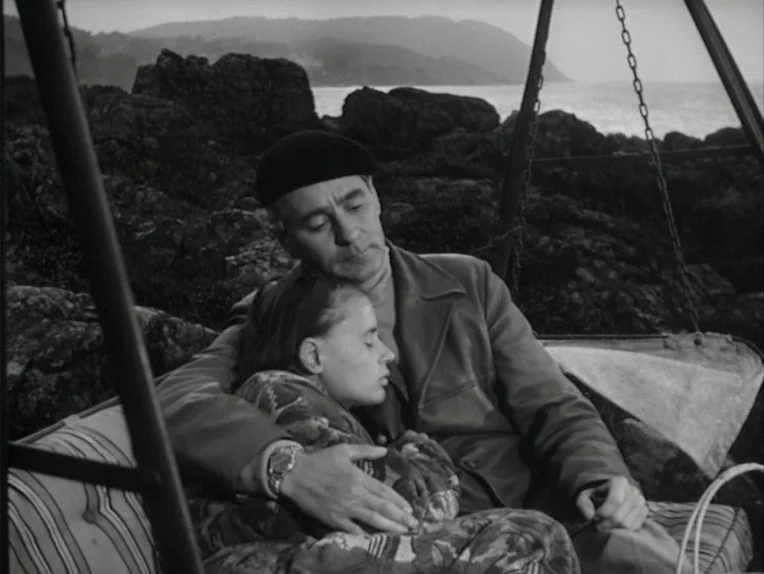 A Lesson In Love (1954) movie still: medium — A man and woman sit together on a swing set overlooking a rocky shore. The background shows a calm…