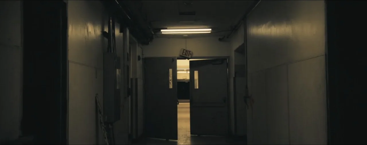 Hunting for Hedonia (2019) movie still: establishing — A long, dimly lit institutional hallway features plain walls and a pair of open doors. The…