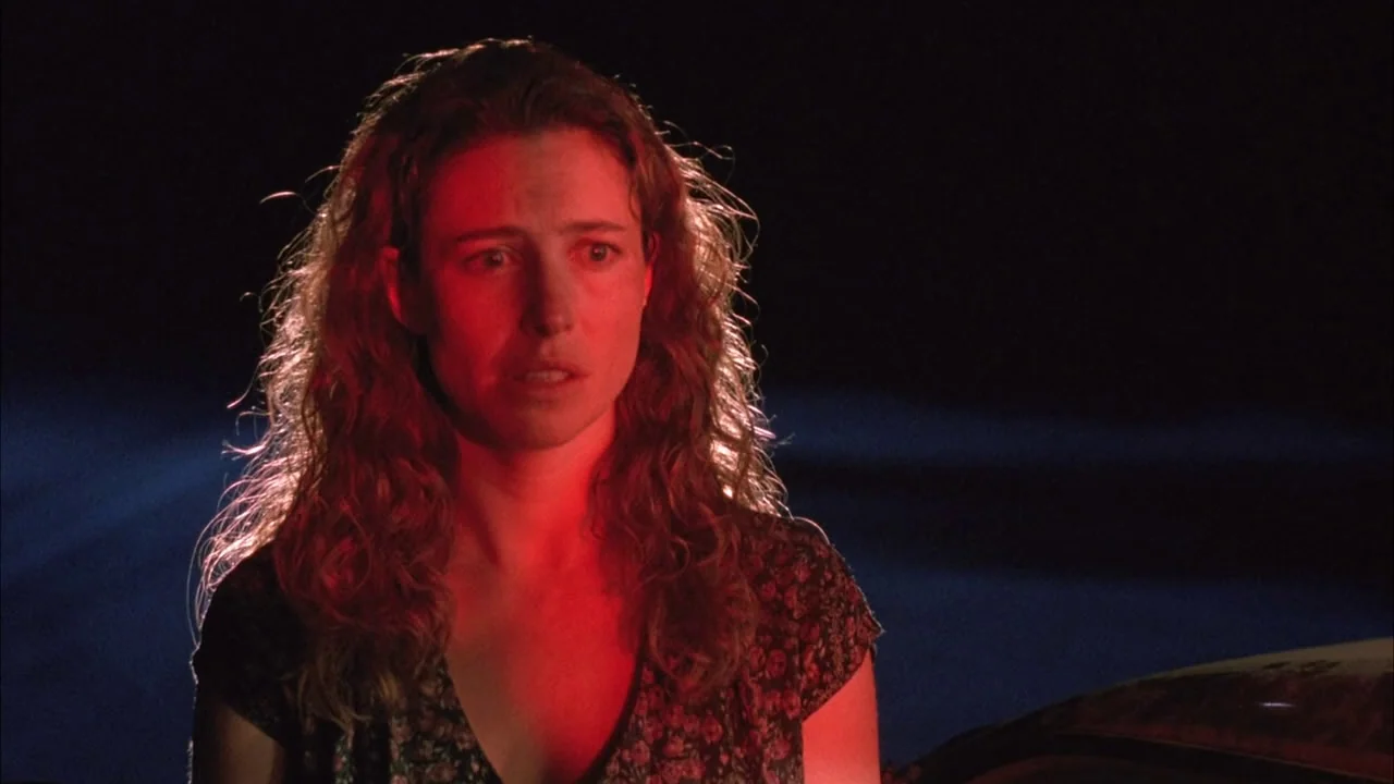 The Rapture (1991) movie still: medium — A woman stands in darkness on the side of a road at night. Red light highlights her face and hair while she…
