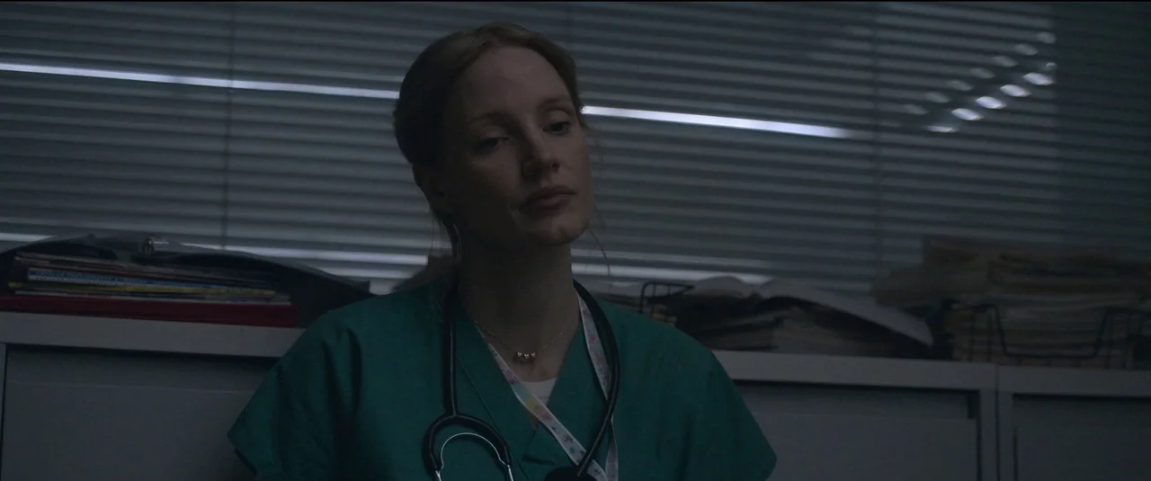 The Good Nurse (2022) movie still: medium — A dimly lit room features a nurse station with stacks of papers and blinds. A woman in green scrubs…