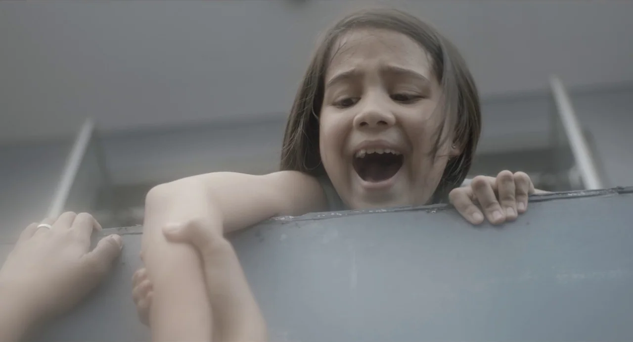May The Devil Take You (2018) movie still: closeup — A low-angle shot shows a child at the edge of a grey ledge. The child screams while an adult's…