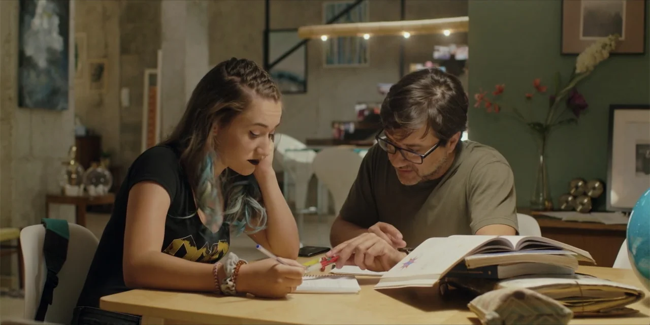 Instant Love (2020) movie still: medium — A modern home dining area featuring a light wood table covered in open books. A man and a young woman sit…