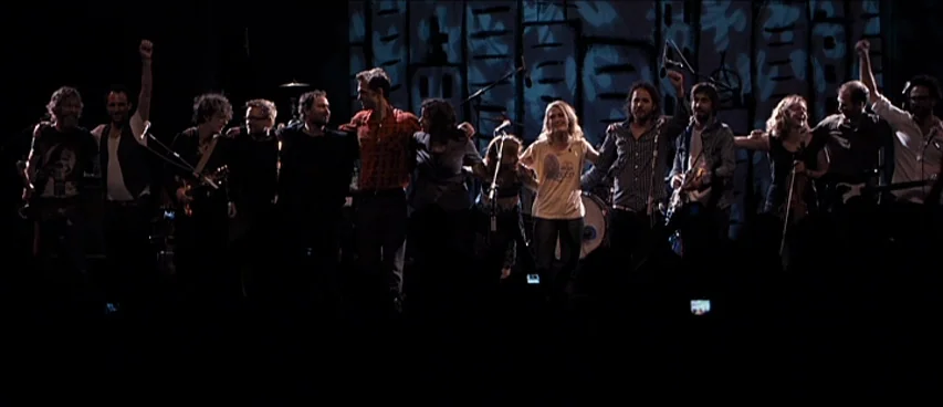 This Movie Is Broken (2010) movie still: wide — A line of musicians stands on a dimly lit stage during a concert performance. The group holds hands…