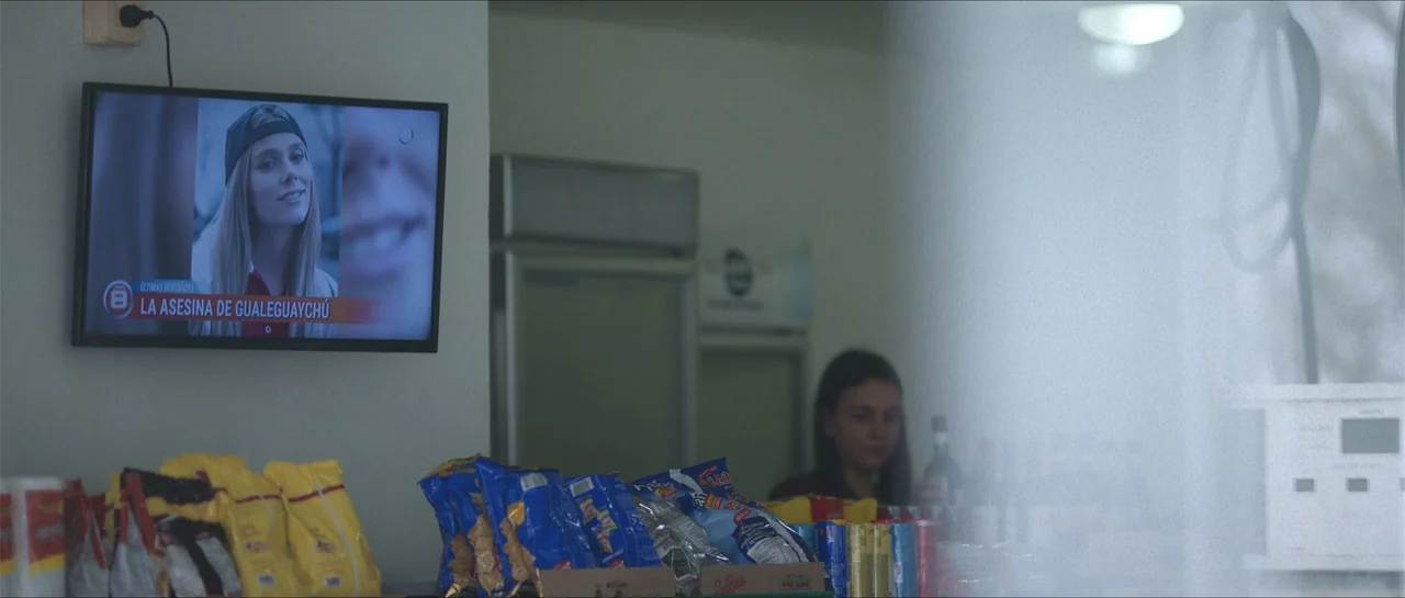 Nahir (2024) movie still: medium — A quiet retail space features a display of snack bags on a service counter. A woman stands in the background as a…
