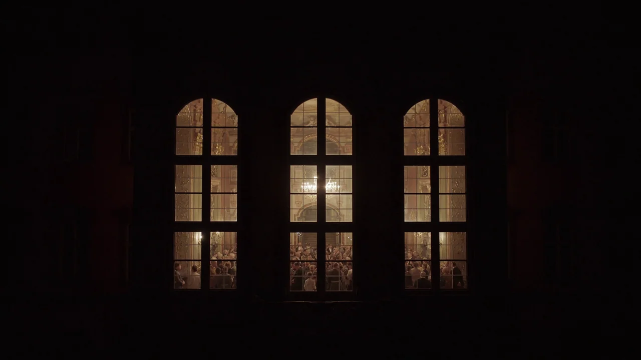 The Zone Of Interest (2023) movie still: wide — Three tall arched windows on a dark exterior wall overlook an brightly lit interior hall. A crowd of…