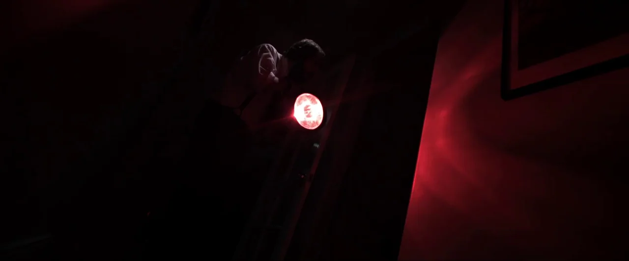 Insidious (2010) movie still: medium — A dimly lit area with dark walls and a singular red light source. A man wearing a white shirt and suspenders…