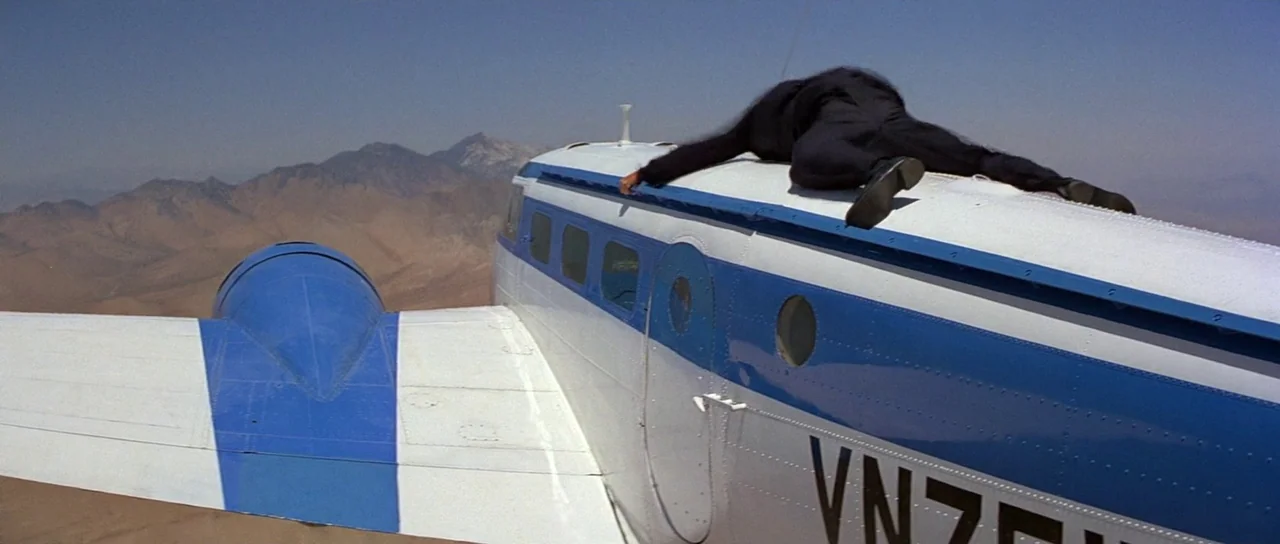 Octopussy (1983) movie still: wide — A man lies prone on the white and blue metal fuselage of a light aircraft. The plane flies over a vast arid…