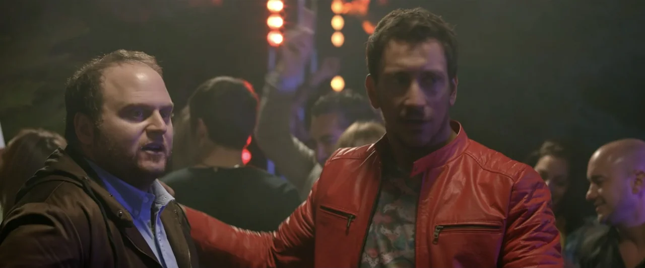 Perdida (2018) movie still: medium — A dimly lit interior space with a blurred crowd and glowing vertical neon lights. Two men face each other in…