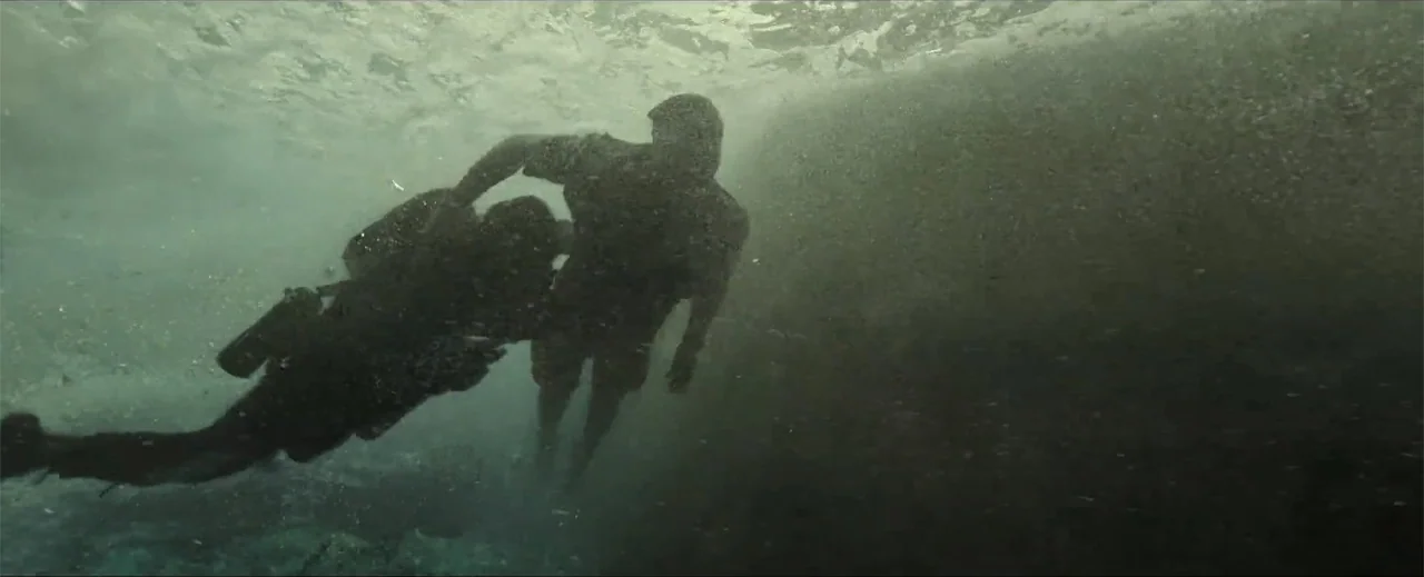 Venom: The Last Dance (2024) movie still: medium — An underwater view of open water with particles floating near a dark rock wall. Two people in…