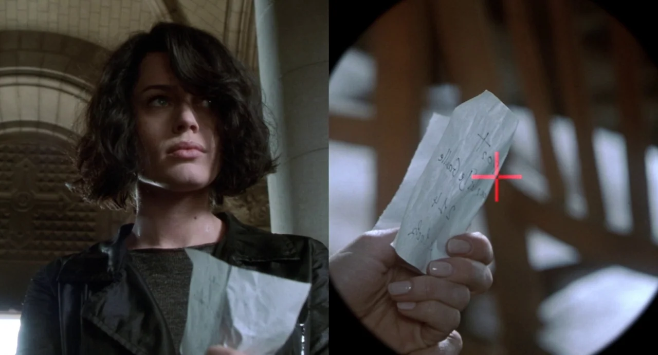 Femme Fatale (2002) movie still: medium — A woman stands in a stone-walled hallway near a large pillar. She holds a small slip of paper that is also…