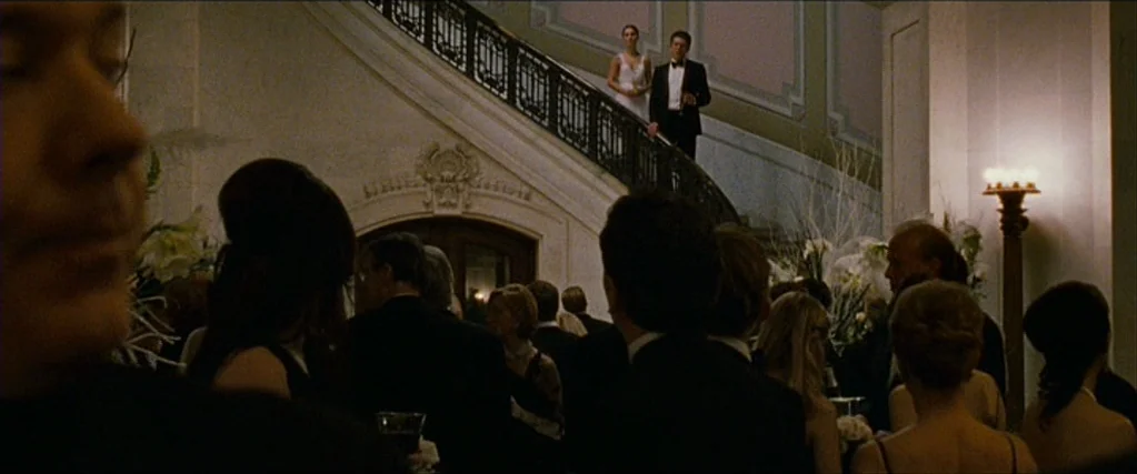 Black Swan (2010) movie still: wide — An opulent ballroom features a grand staircase with an ornate iron railing. A couple walks down the stairs…