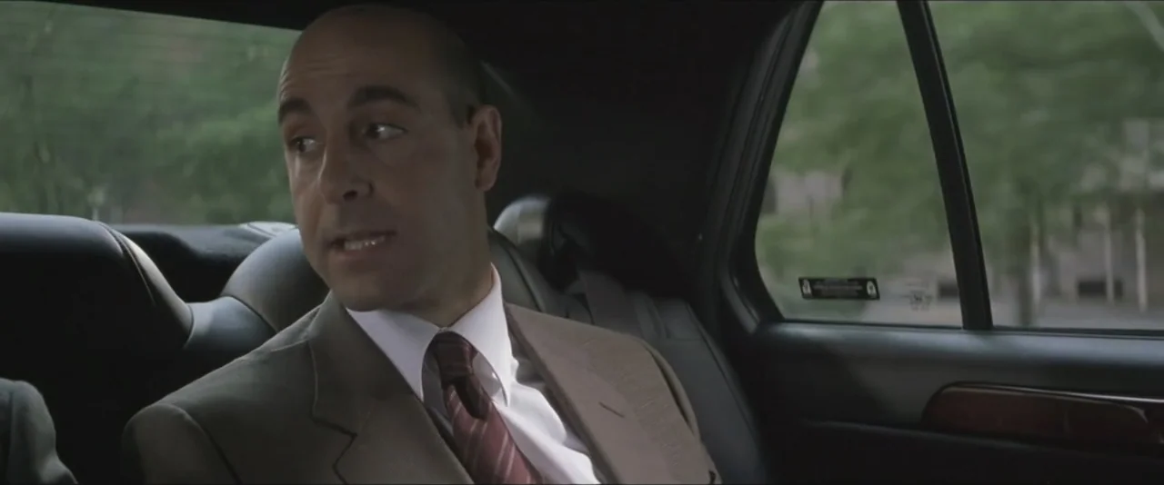 Maid in Manhattan (2002) movie still: medium — A man sits in the backseat of a moving vehicle with visible trees outside. He wears a formal suit and…