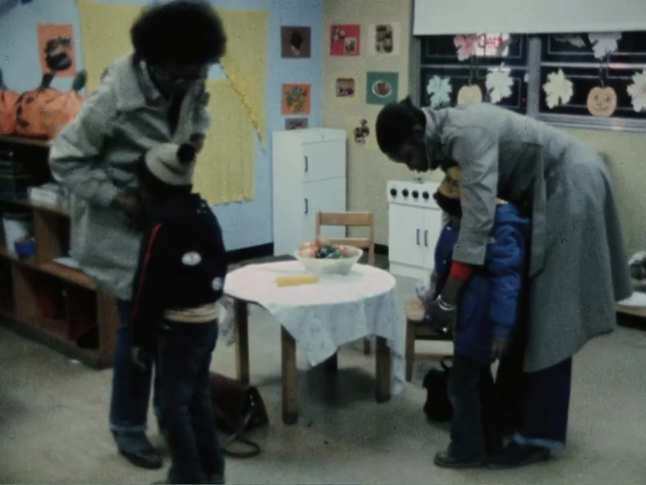 Born in Flames (1983) movie still: medium — A 1970s school room features festive paper decorations and a central wooden table. Two women stand in…