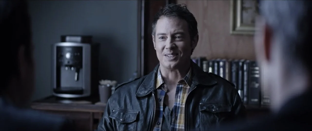 Room 6 (2006) movie still: medium — A dimly lit room features a bookshelf and a coffee machine in the background. A man in a leather jacket smiles…
