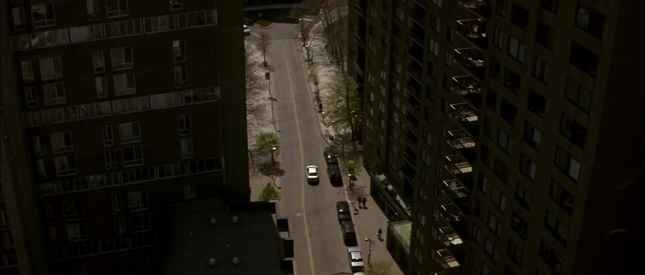 Dark Water (2005) movie still: establishing — An urban canyon formed by two tall apartment buildings borders a narrow paved road. A single white car…