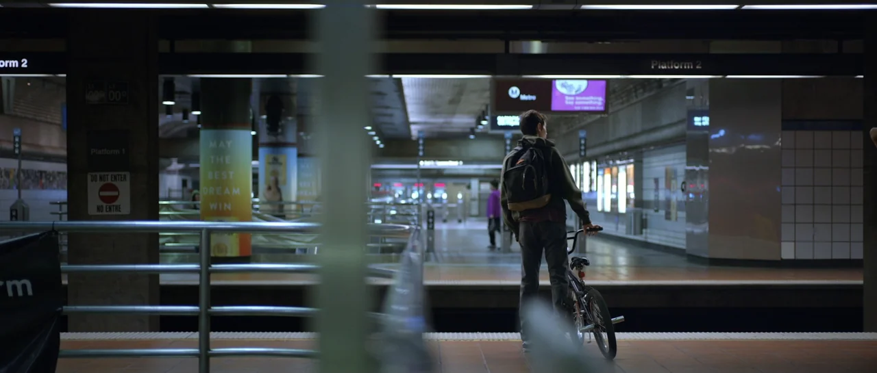 La presencia (2012) movie still: fullbody — A quiet, modern subway station with orange structural pillars and clean tiled floors. A young man with a…