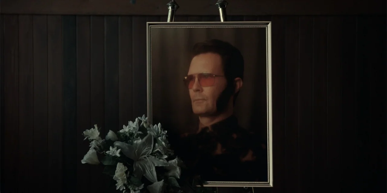 The Monkey (2025) movie still: medium — A framed portrait of a man sits on an easel against dark wooden paneling. A small bouquet of white lilies…
