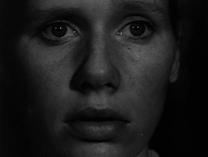 Hour of the Wolf (1968) movie still: closeup — A close portrait of a person is set against a featureless black backdrop. The individual gazes…