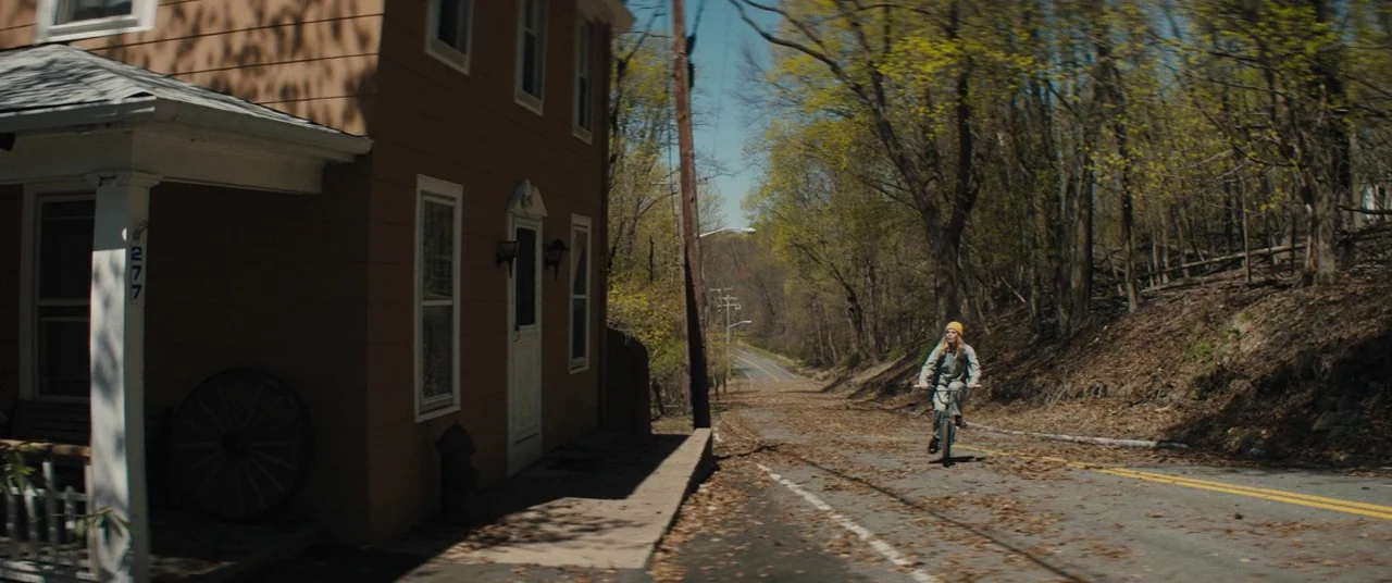 I Think We’re Alone Now (2018) movie still: wide — A quiet rural road passes by a wooden residential house with a covered porch. A woman rides a…