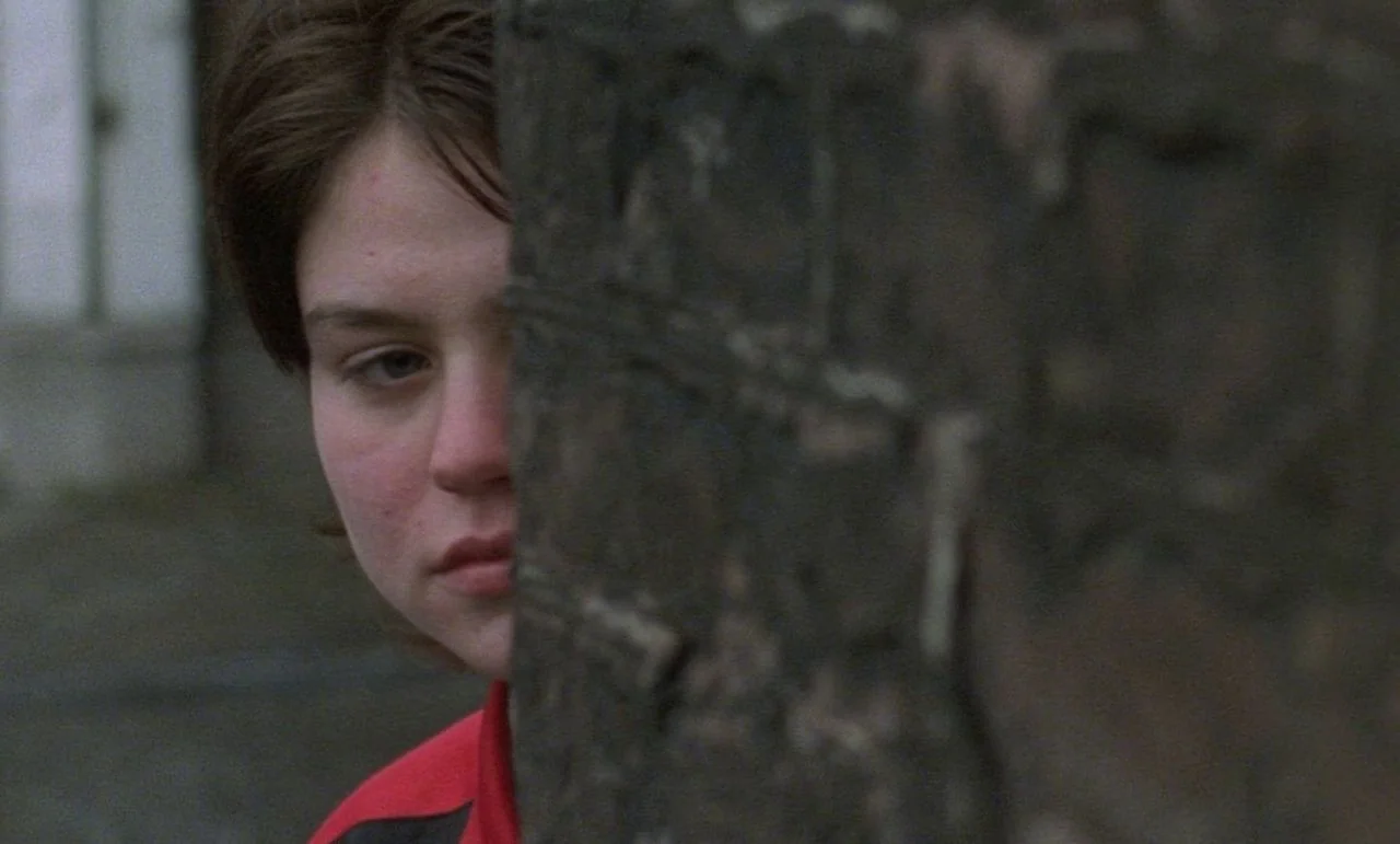 Rosetta (1999) movie still: closeup — A teenager stands behind a dark, textured concrete post in a dim urban setting. The girl looks past the edge…
