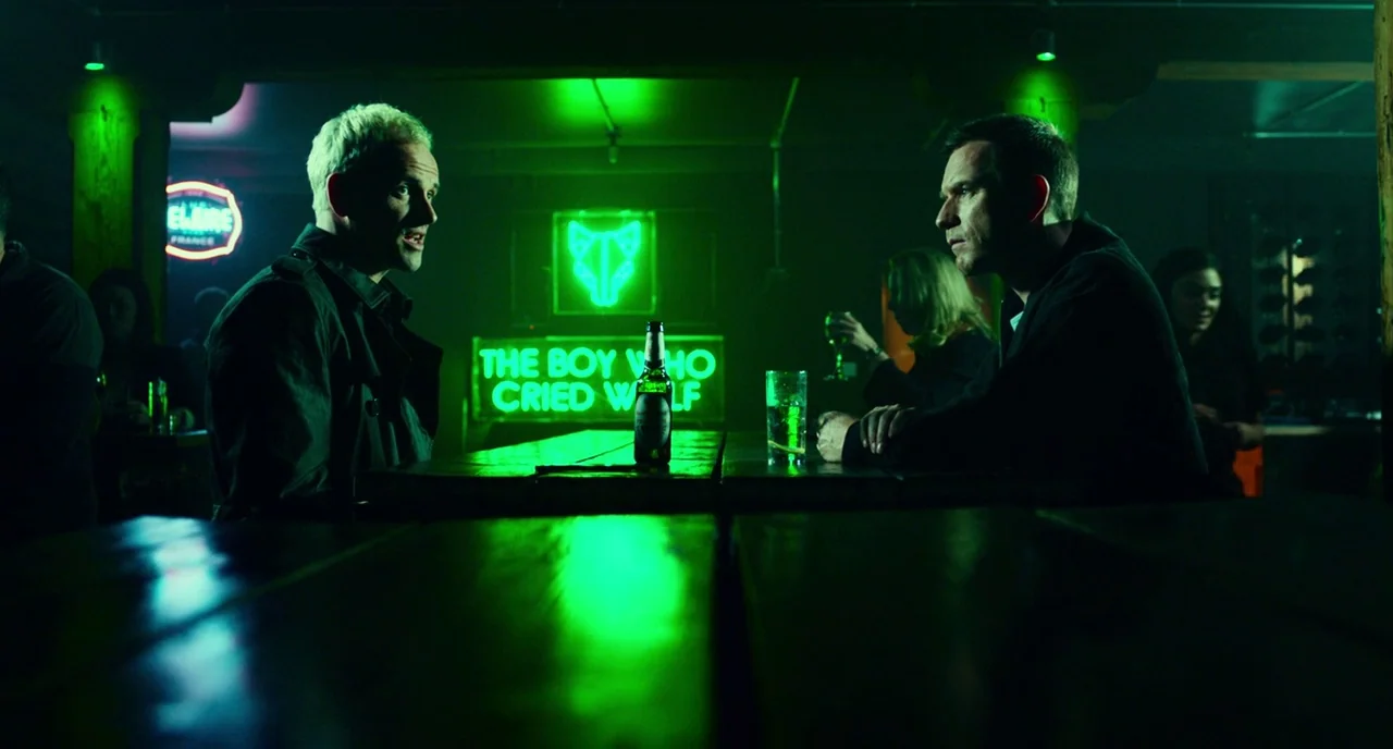 T2 Trainspotting (2017) movie still: medium — A dimly lit room is illuminated by bright green neon signs against the wall. Two men sit at a long…
