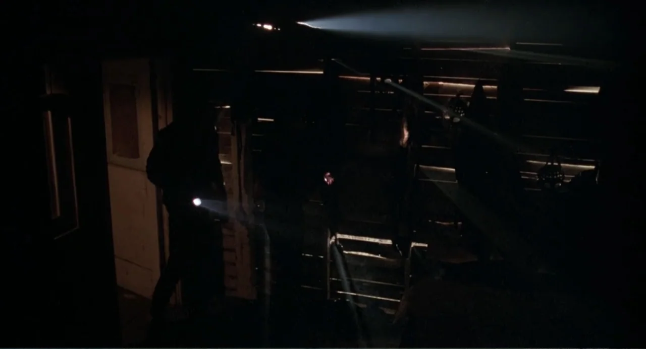 Dangerously Close (1986) movie still: medium — A dark, wooden-walled cabin interior is illuminated by narrow beams of artificial light. Two figures…