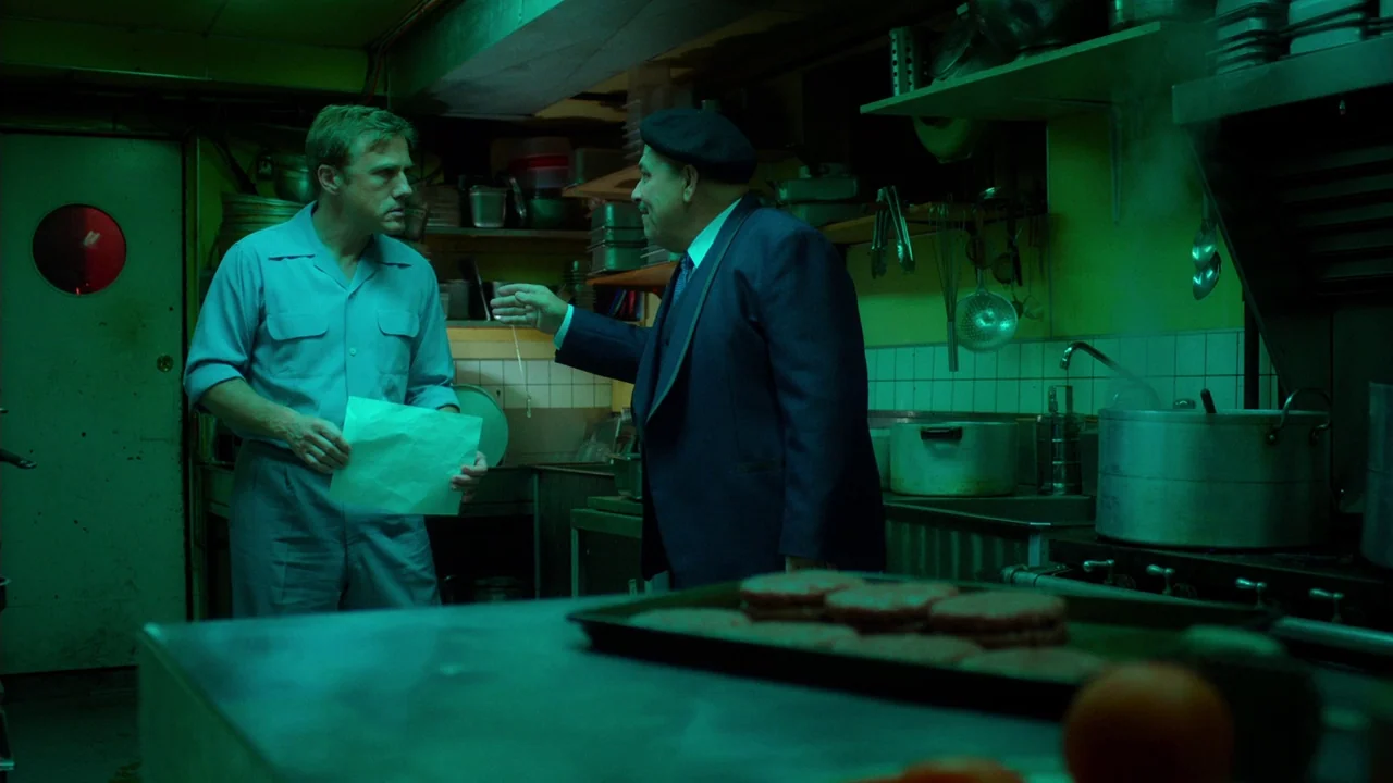 Big Eyes (2014) movie still: medium — A dark restaurant kitchen is bathed in a distinct green light. Two men stand near a stainless steel counter…