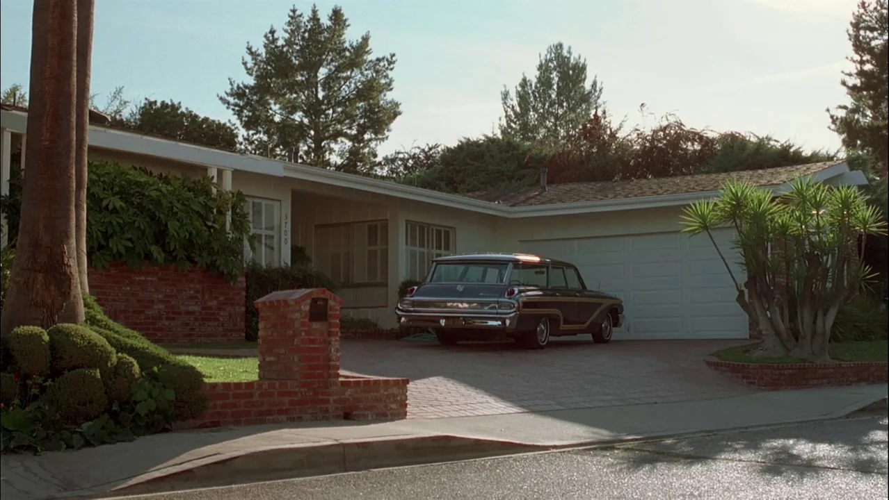Auto Focus movie still: wide — A 1960s single-story suburban home features brick accents and a pale facade. A vintage dark station wagon is parked…