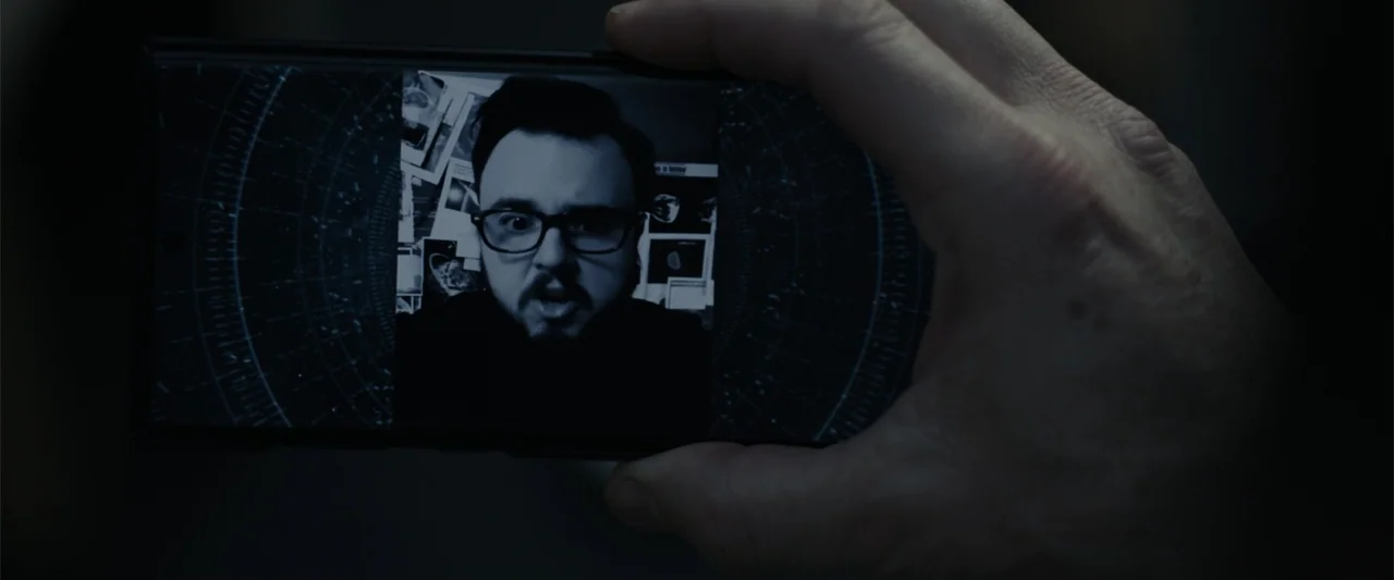 Moonfall (2022) movie still: closeup — A smartphone screen displays a video feed of a man within a dimly lit room. The hand of an unseen person…