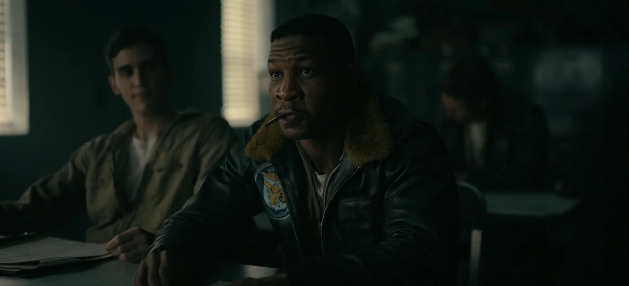 Devotion (2022) movie still: medium — A dimly lit classroom features rows of desks with windows covered by slatted blinds. A man in a leather flight…