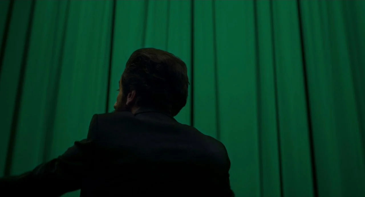 Annette (2021) movie still: medium — A man in a dark suit stands before a floor-to-ceiling green curtain. He faces away from the camera, looking…