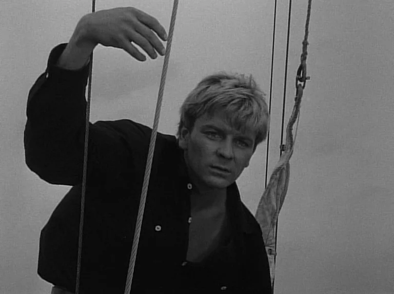 Knife In The Water (1962) movie still: medium — A man stands on a sailboat deck surrounded by thin vertical tensioned cables. The man holds a…