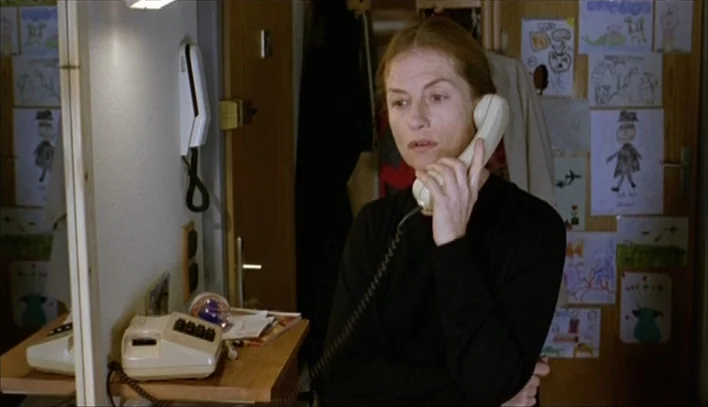 The Piano Teacher (2001) movie still: medium — An interior hallway has a light-colored wooden door covered in several children's drawings. A…