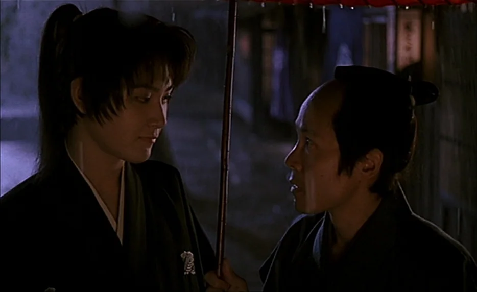 Gohatto (1999) movie still: medium — Two individuals stand in a dark, wet environment illuminated by faint night light. One holds a red paper…