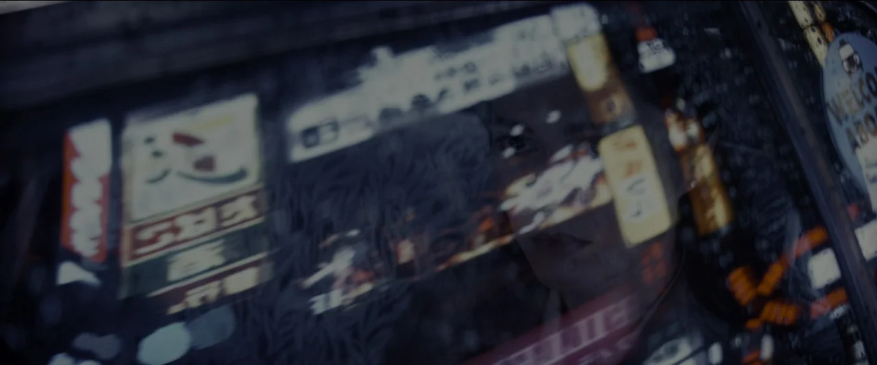 Kate (2021) movie still: closeup — A dark night street scene viewed through a store window covered in advertisements. A person's face is partially…