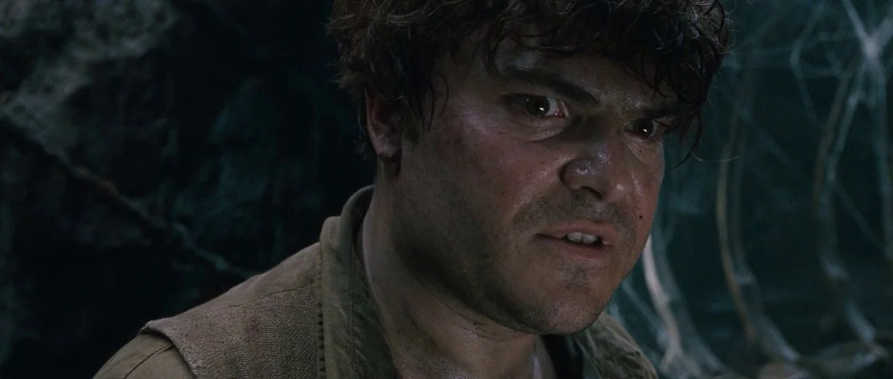 King Kong (2005) movie still: closeup — A dark, rocky cave interior is filled with shadows and faint web-like textures. A man with disheveled hair…