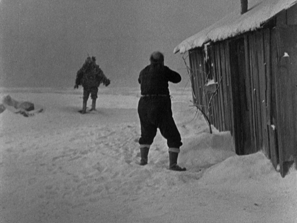 The Gold Rush (1925) movie still: wide — A snow-covered landscape features a small wooden structure with a snow-laden slanted roof. A man in dark…