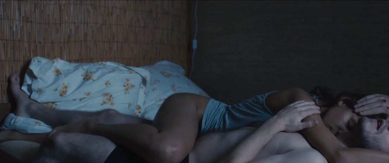 Simon Killer movie still: medium — A dimly lit bedroom features a bamboo wall covering behind a bed. A couple sleeps intertwined under white linens…