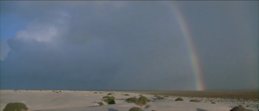 Roadgames (1981) movie still: establishing — A vast, flat desert landscape stretches under a heavy, dark overcast sky. A single rainbow arcs down…