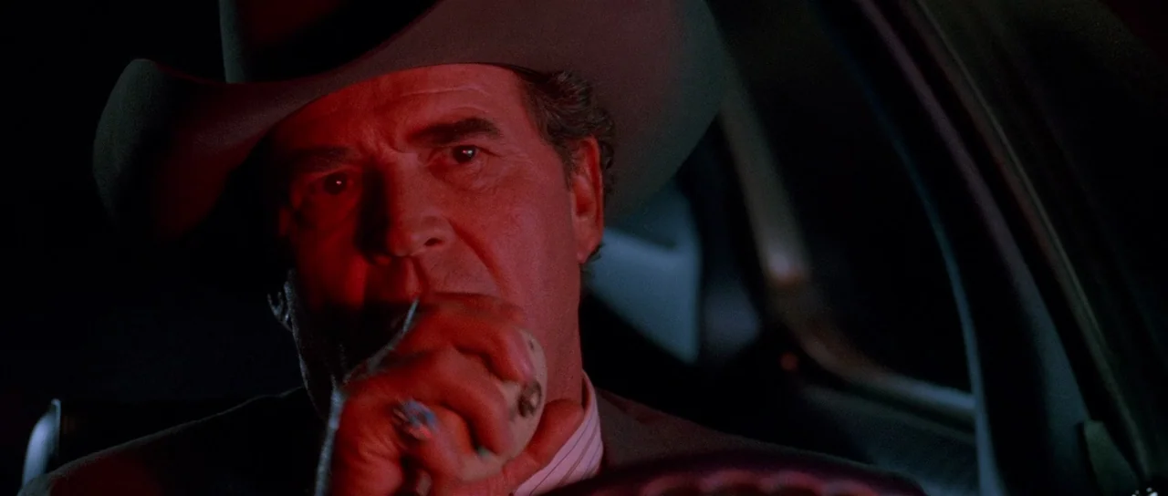 Fire In The Sky (1993) movie still: closeup — The interior of a vehicle at night is bathed in a harsh red light. A man wearing a cowboy hat sits in…