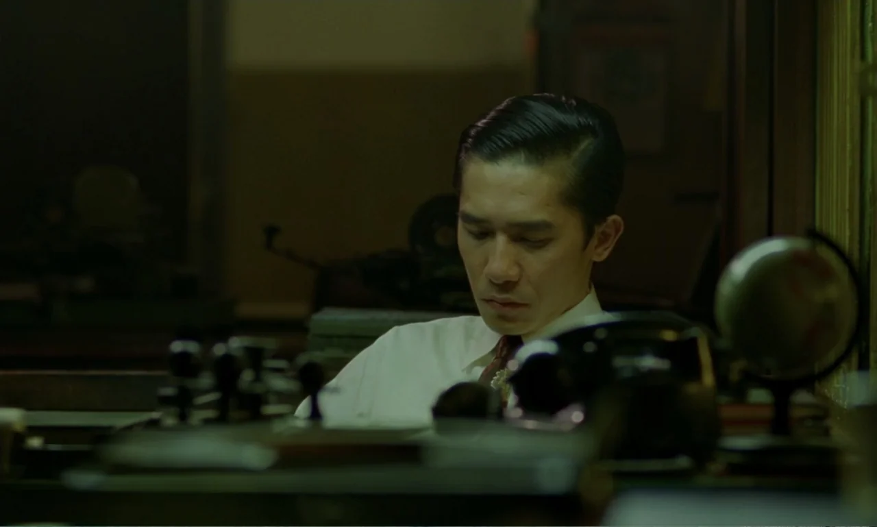 In the Mood for Love (2000) movie still: medium — An dimly lit office features vintage equipment and a globe on a desk. A man in a dress shirt sits…