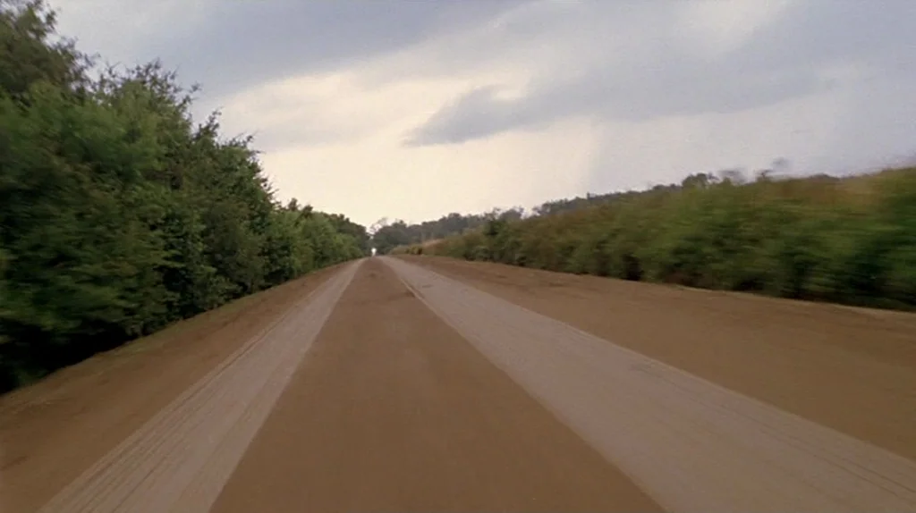 Undertow (2004) movie still: establishing — A long, unpaved dirt road flanked by dense green foliage on both sides. Cloudy sky hangs over the…