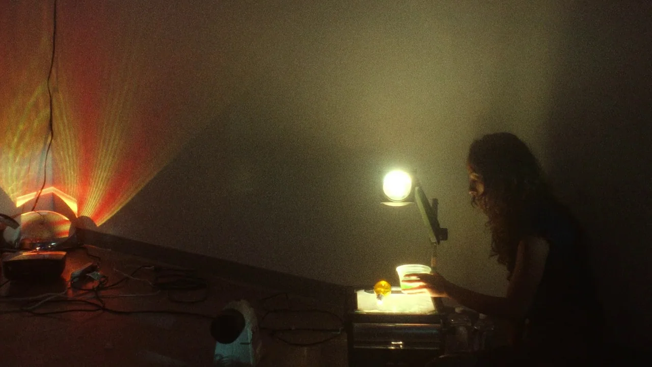 Showing Up (2022) movie still: medium — A dimly lit room with walls catching projected colored light patterns from electronic devices. A young woman…