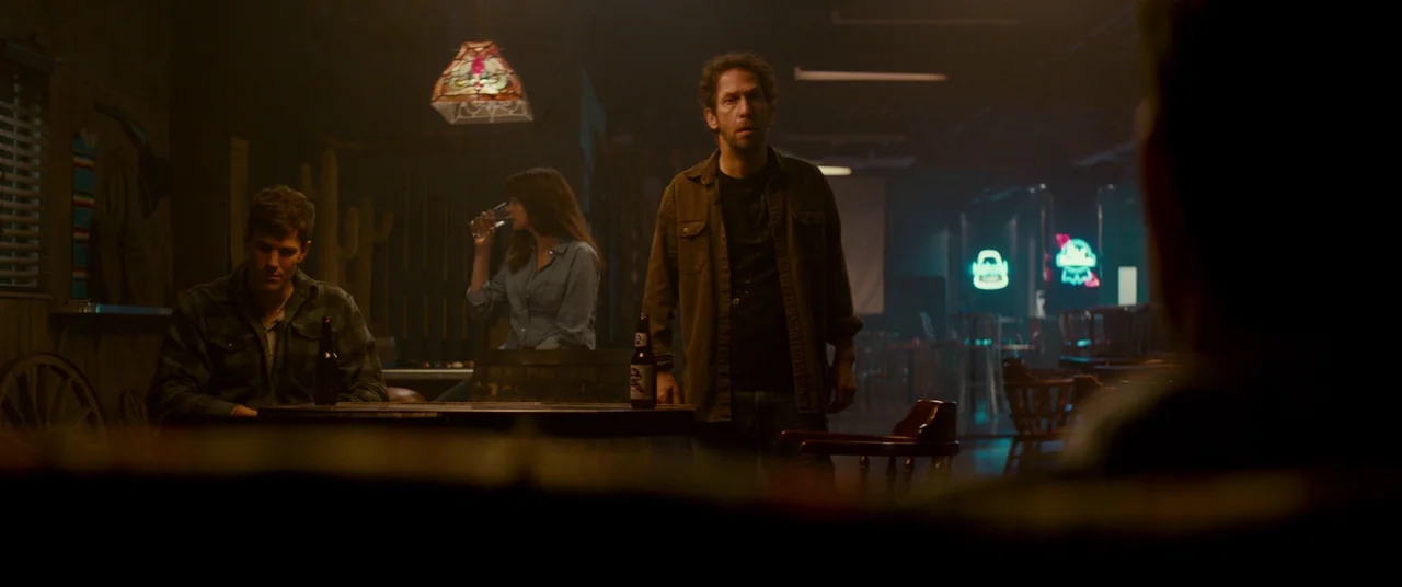 Colossal (2016) movie still: wide — A dark, wood-paneled bar featuring neon beer signs and hanging stained glass lamps. Three patrons occupy tables…