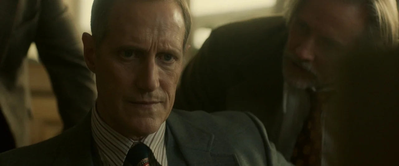 Stockholm (2019) movie still: closeup — An interior space with muted colors contains two men in professional business attire. The primary subject…